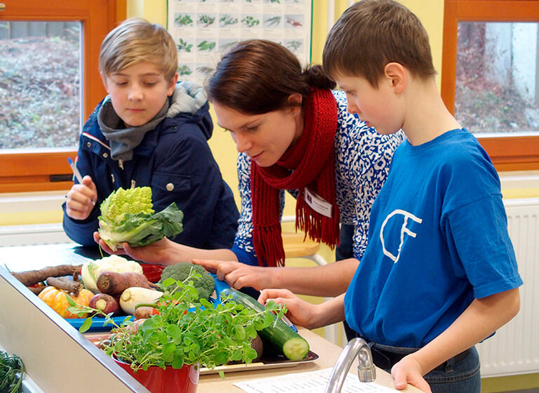 Lehrerin klärt zwei Schüler über verschiedene Gemüsearten, die vor Ihnen liegen, auf.