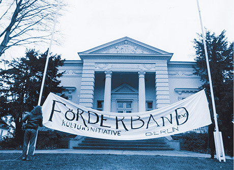 Weißes Banner aufgehangen zwischen zwei Fahnenmasten vor weißem Gebäude mit dem Schriftzug Förderband e.V. Kulturinitiative Berlin
