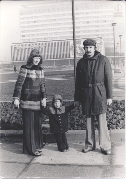 Auf einer alten Schwarz-Weiß-Fotografie steht ein Paar mit kleinem Kind in 70er Jahre Kleidung vor DDR-Architektur.