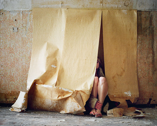 Eine junge Frau mit nackten Beinen und Füßen sitzt halb verdeckt hinter von der Wand herab gerollten alten Tapetenbahnen.
