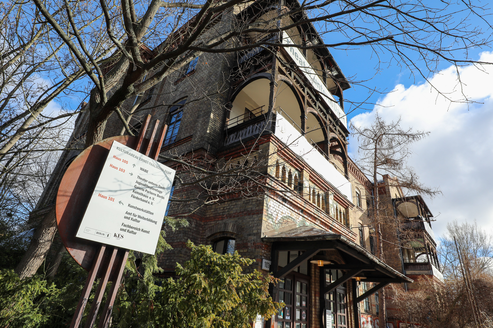 Vor einer mehrstöckigen historischen Backsteinvilla mit verzierten Holzbalkonen steht ein Schild: Kulturarreal Ernst-Thälmann-Park