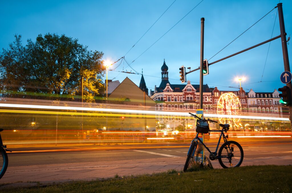 Hinter dem Lichtstreifen eines schnell durchfahrenden Zuges zeichnen sich mehrstöckige Gründerzeithäuser und ein hell beleuchtetes Riesenrad ab.