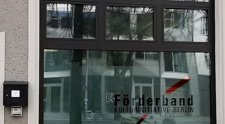 Auf einer großen Frontscheibe spiegeln sich Bäume und die Fensterfront eines gegenüber liegenden Hauses. Auf der Scheibe prangt das Förderband Logo- Links neben der Scheibe hängt ein Briefkasten, darunter eine Klingel.