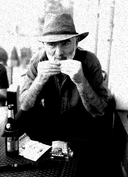 Schwarz-Weiß-Foto. Ein älterer Herr mit Strohhut und grauem Vollbart sitzt an einem Kneipentisch, leckt eine selbstgedrehte Zigarette an und lächelt verschmitzt in die Kamera.