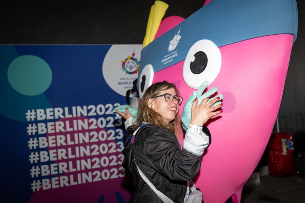 Eine lachende junge Frau mit Brille umarmt das herzförmige rosa Maskottchen der Special Olympics. Im Hintergrund steht mehrfach untereinander #Berlin 2023